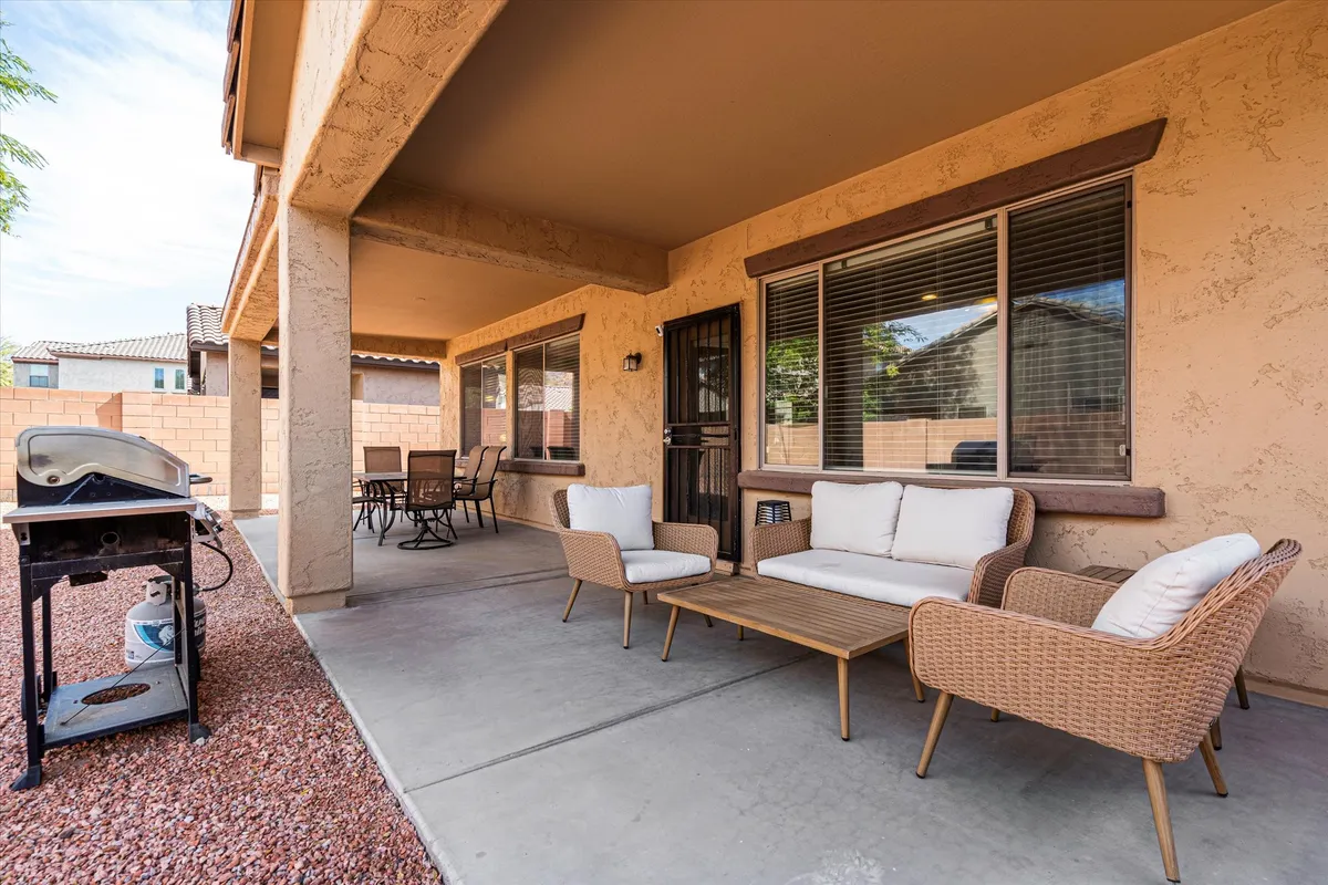 A comfortable backyard patio area featuring wicker seating, an outdoor dining set, and a grill. The scene is bathed in warm light, suggesting the approach of sunset, and the space feels private and ready for gatherings or relaxation.