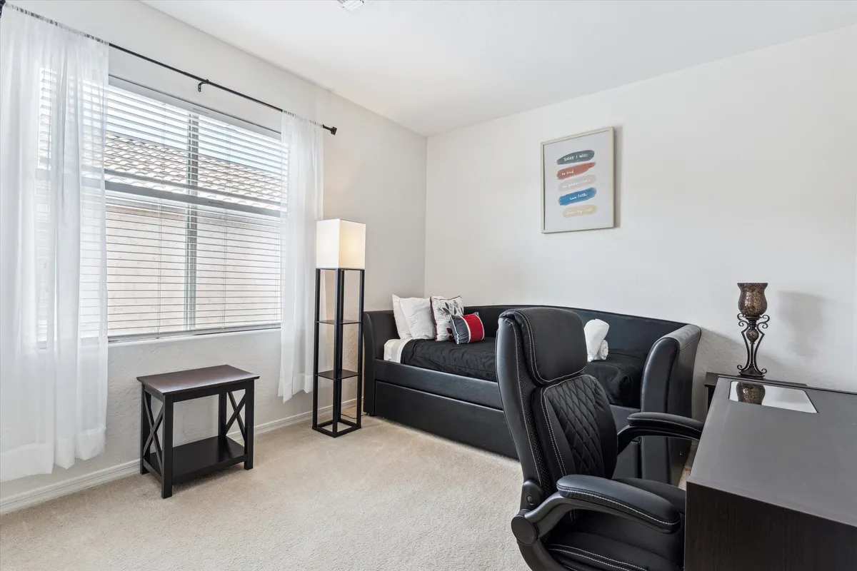 A bedroom with a daybed and plush pillows, a dedicated desk and chair, and a window with sheer curtains allowing soft daylight to fill the room. The space is designed for both rest and productivity, with a cozy atmosphere ideal for working or unwinding.