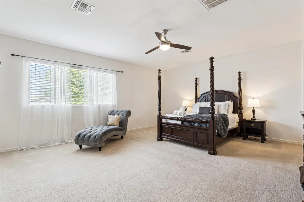 A serene primary bedroom suite featuring a four-poster bed adorned with luxurious bedding, a chaise lounge for relaxing, elegant nightstands with lamps, and ample natural light streaming in. The overall atmosphere is tranquil and inviting, ideal for restful nights and rejuvenating mornings.